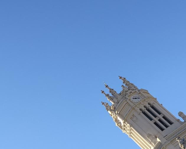 Clock tower with ornate spires against a clear blue sky. The tower is an off-white or cream color with detailed Gothic-style architectural elements, including pointed pinnacles and carved decorations. Near the top is a round clock face with black hands and markings, showing the time as about 4:20. Below the clock, there are tall, narrow windows with dark openings. The photo is taken at an angle so the tower slants from the lower right toward the upper center, and the rest of the image is open sky.