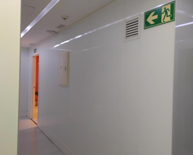 White hallway in a modern building. The corridor has smooth white walls and a white ceiling with recessed lighting running along it. On the right upper part of the image, there is a green emergency exit sign with a running figure and a large left-pointing arrow, indicating the exit is toward the left. Just to the left of that sign is a rectangular wall vent. Farther down the hallway on the left side, there is an open doorway; inside that room the walls or lighting appear orange, and a darker object is partl