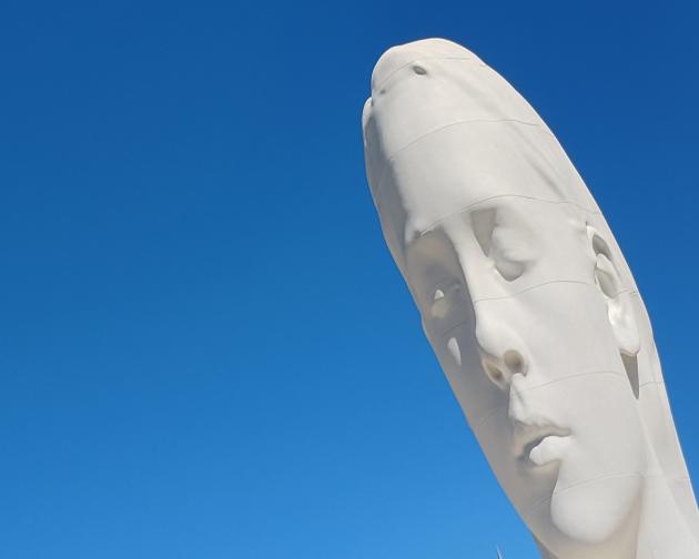 A tall, elongated white sculpture of a human face against a clear, bright blue sky. The face is stylized and stretched vertically, with soft, smooth features and closed eyes, giving a calm, meditative expression. Subtle horizontal bands or seams wrap around the surface, as if the sculpture is made from stacked or slightly shifted layers. The head is tilted gently to one side, and only the upper part of the sculpture is visible in the frame, with no other objects or background elements present.