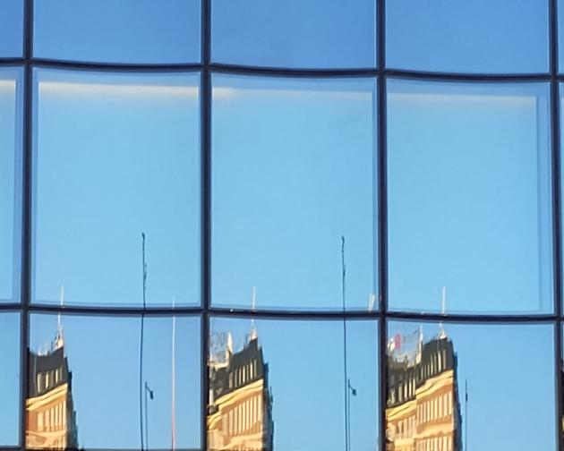 Blue-tinted glass windows of a modern building reflecting an older city building. The surface is divided into a grid of tall rectangular window panes with slightly curved frames, creating a repeating pattern. In the lower row of panes, the reflection of a traditional multi-story building appears, with warm yellow-orange walls, white window frames, and a dark roof. The reflected building is stretched and slightly distorted differently in each pane, so its roofline rises and falls in a wavy rhythm across it.