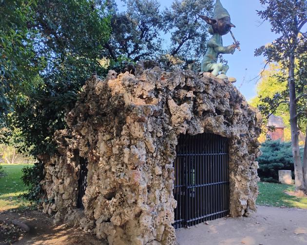 Rocky grotto-like structure in a park setting. The building is made of rough, irregular stones, forming a rounded, cave-shaped hut. The front has a large arched entrance closed off with black vertical metal bars, like a gate, and there is a smaller barred opening on the left side. On top of the rocky roof, there is a greenish statue of a small fantasy creature resembling a goblin or elf, sitting cross-legged and playing a flute; it has a pointed hat, long pointed ears, and exaggerated facial features. 