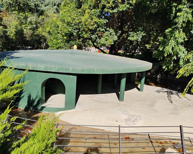 Low, round, green concrete shelter structure in an outdoor setting. The building has a flat circular roof supported by several cylindrical green pillars at the front. On the left side of the structure there is an arched doorway opening into a shaded interior space. The ground around the shelter is light-colored concrete, with a shallow channel or water area in front that has a few scattered leaves and some rocks. In the foreground, a short black metal railing runs horizontally. 