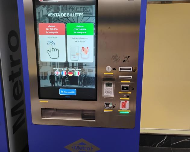 This is a blue Metro ticket machine with a large touch screen in the center. Above the screen, from left to right: Card slot for bank cards, Cash note slot, Contactless card area, Ticket/card slot with a green light, Keypad with a small display, Ticket/receipt output slot. On the screen you can choose ticket options and languages, and there is an accessible-use button with a wheelchair symbol. Summary provided by Be My Eyes AI.