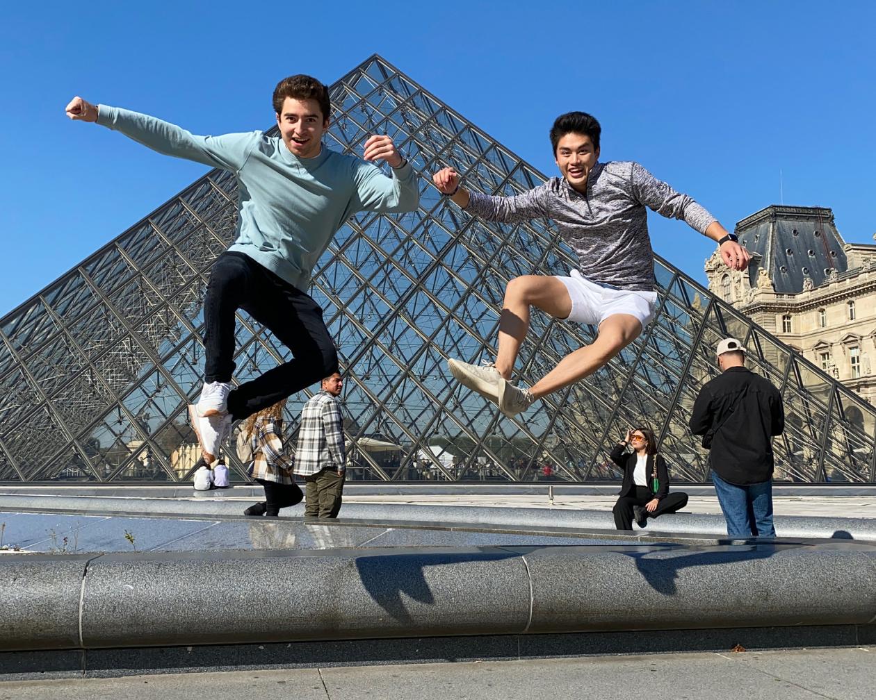 Two students are jumping and smiling in front of the Louvre Pyramid in Paris, France.