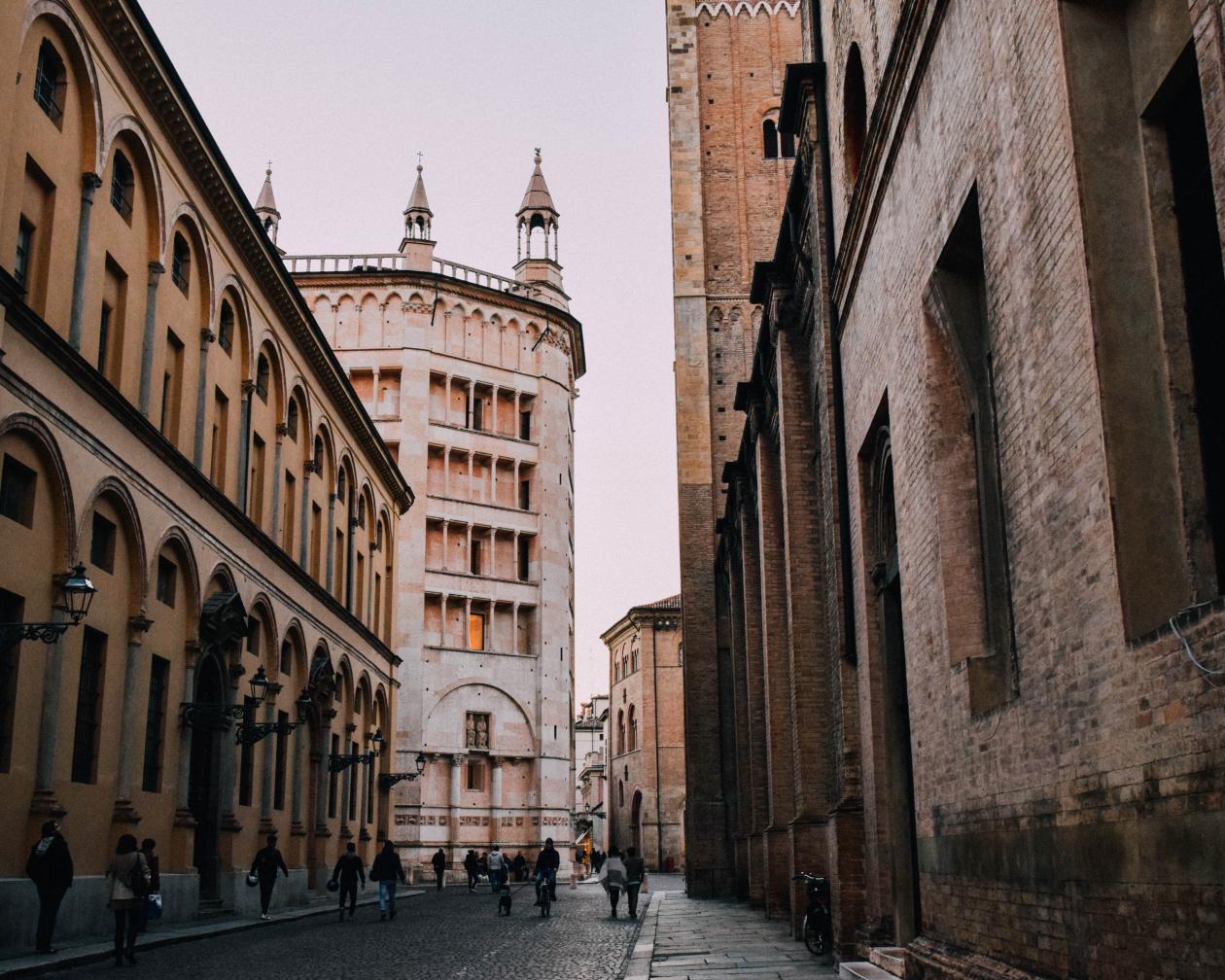 People walk in the historic center of Parma. The buildings are made of red brick and some have spires atop them.