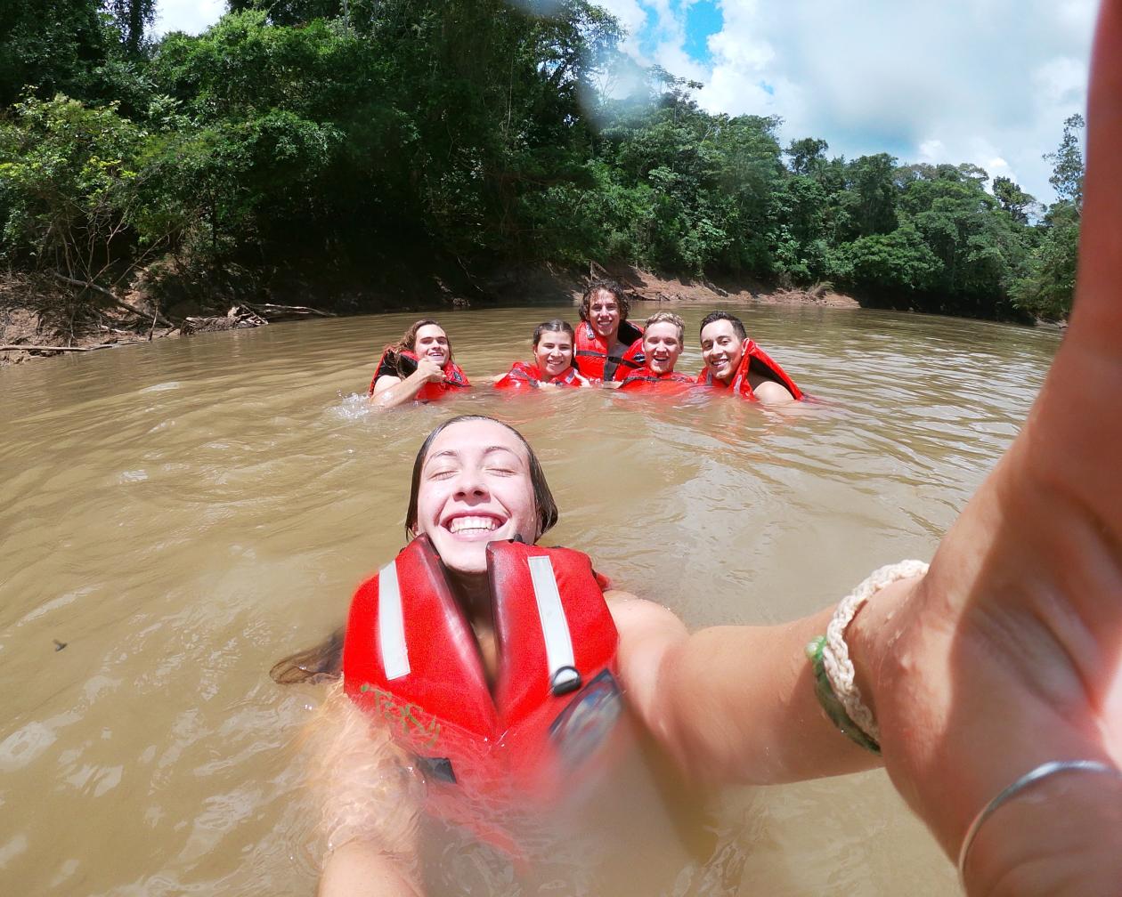 One student taking a selfie while swimming in the Amazon Rainforest with a group of other students in the distance behind her