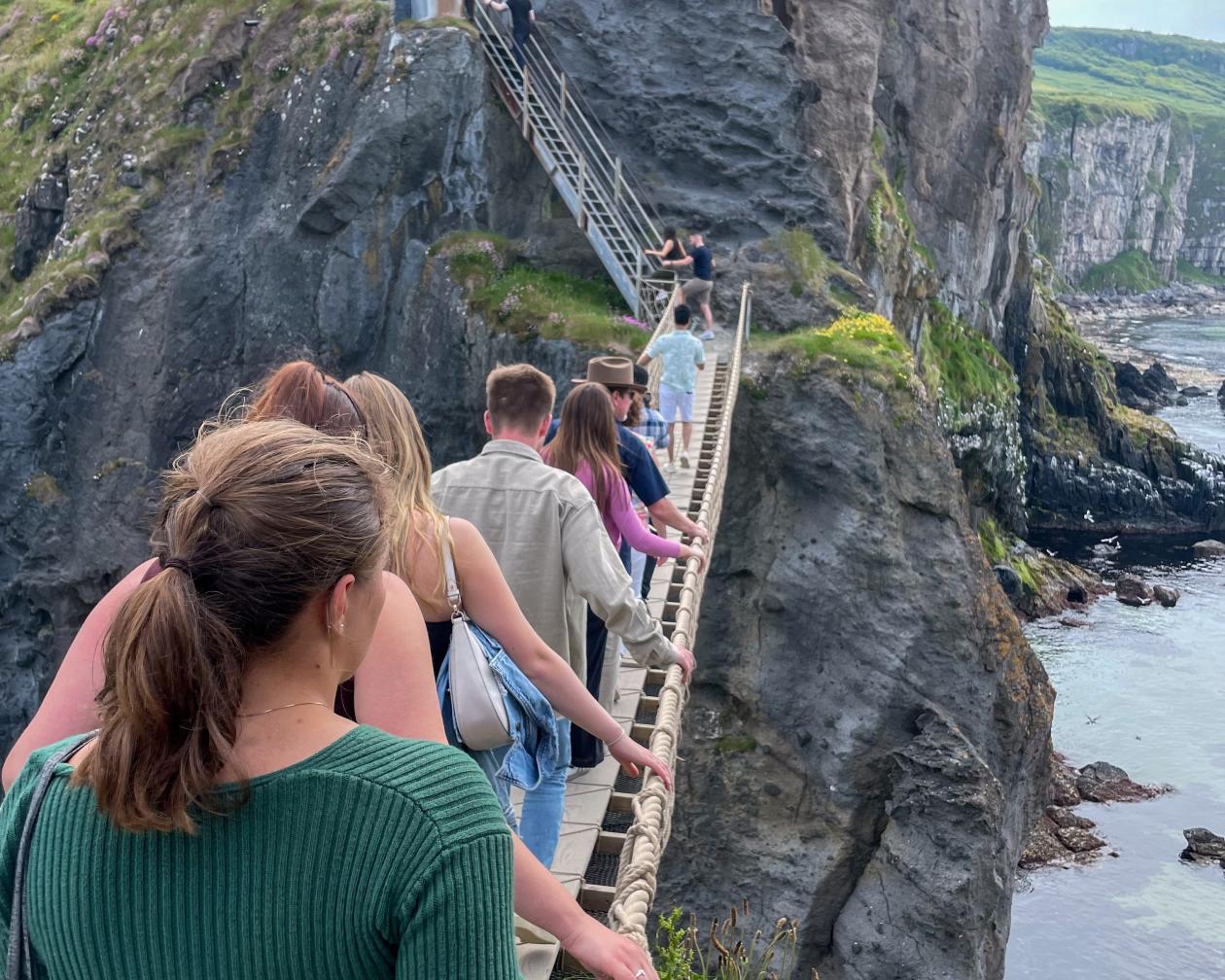 line of students walking on a rope bridge across an oceanside mountanous terrain