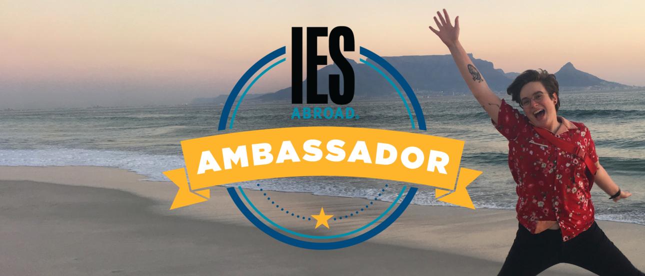 a student on a beach waving at the camera behind the IES Abroad Student Ambassador logo
