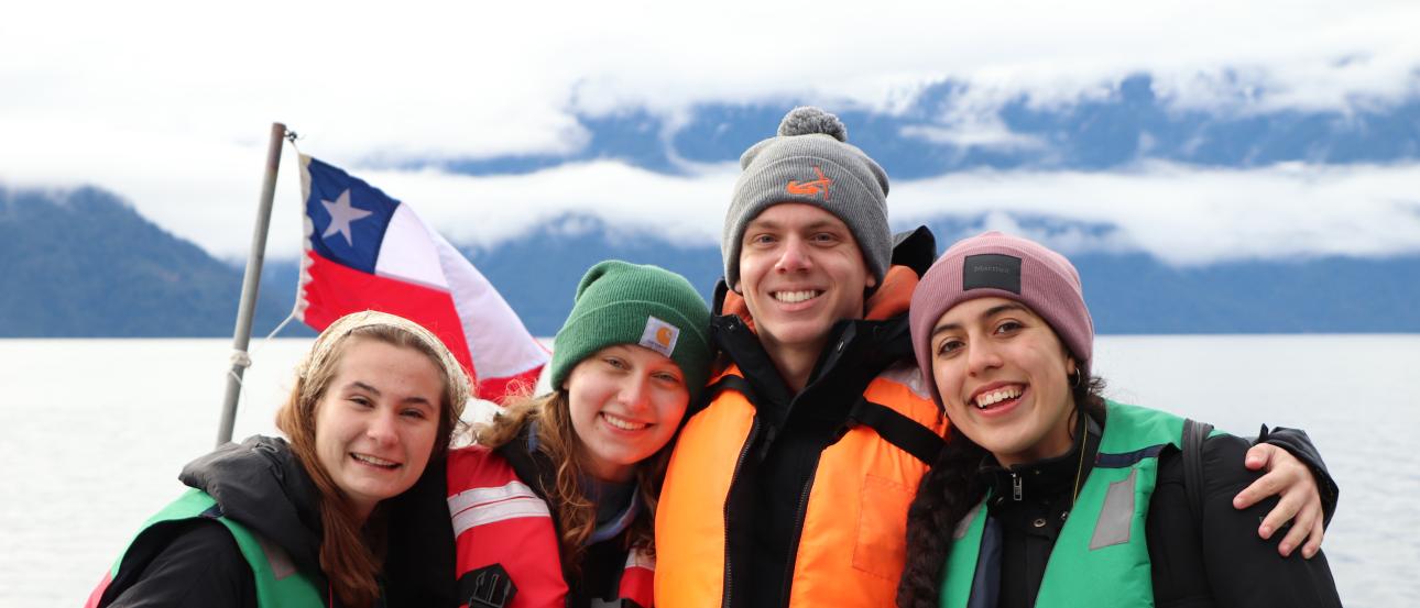 Four students wearing red, orange, and green multi-colored life vests are on a boat in Santiago, Chile.