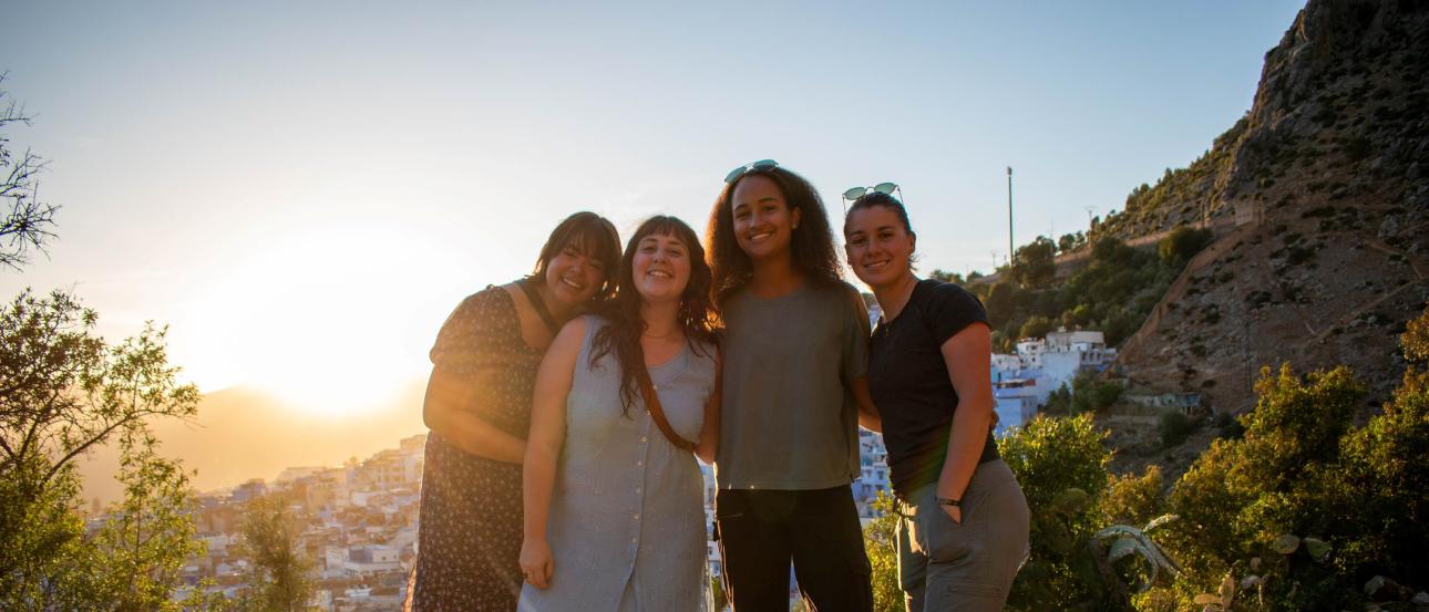 four students standing on grass and smiling with a mountanous sunset in the background
