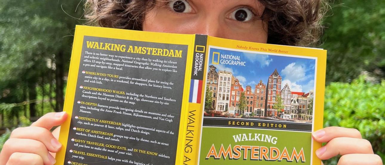 A guidebook titled "Walking Amsterdam" is being held open by Brooke, a white woman with brown curly hair, whose eyes are looking over the top of the book at the camera. 