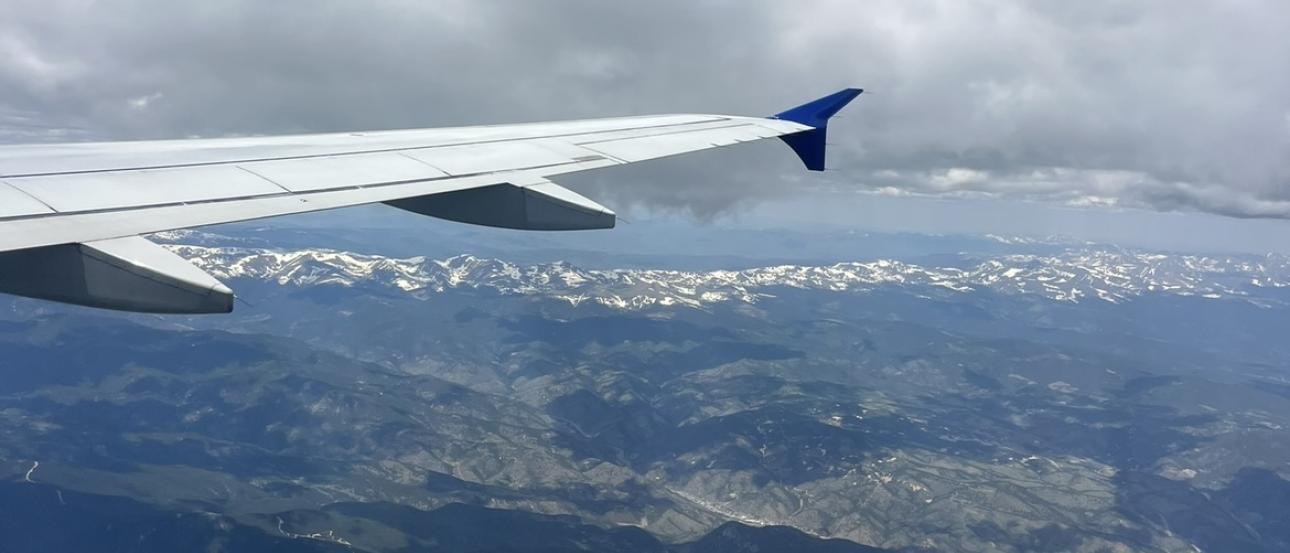 The right wing of a plane cuts across the sky, as viewed from the window seat. Cloudy blue skies look down on a shadowy mountain terrain.