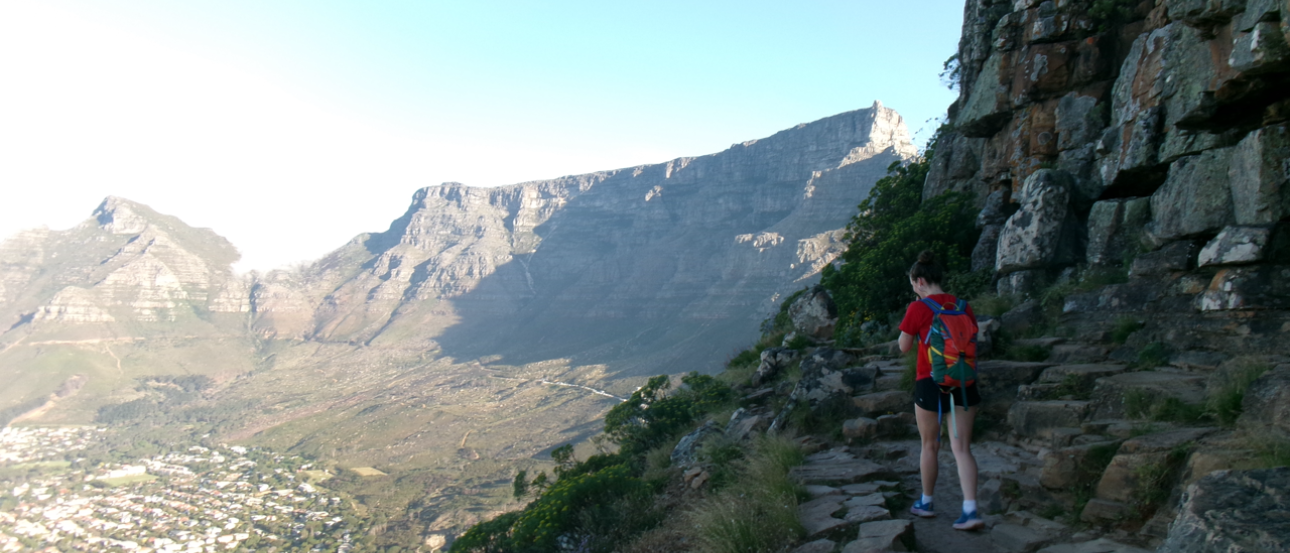 The view of Table Mountain from Lion's head. On the right there is a person walking and a rocky terrain. 
