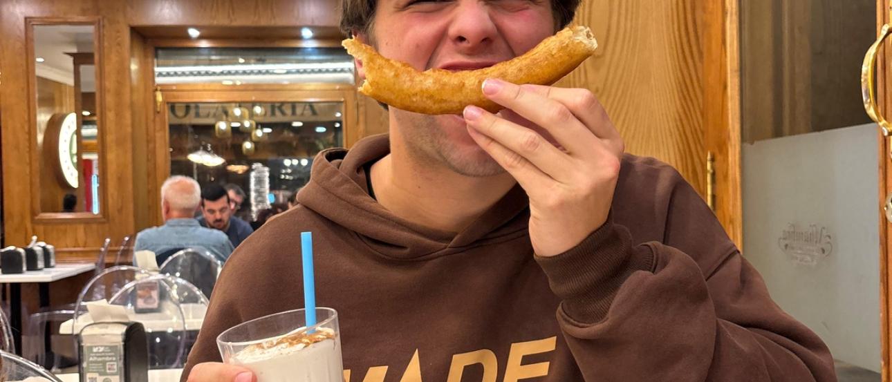 My friend Caden sits inside the churro shop at night, smiling. His face is scrunched up and he holds a curved churro in front of his mouth to mimic a smile, clutching a cinnamon-dusted leche rizada in the other hand.