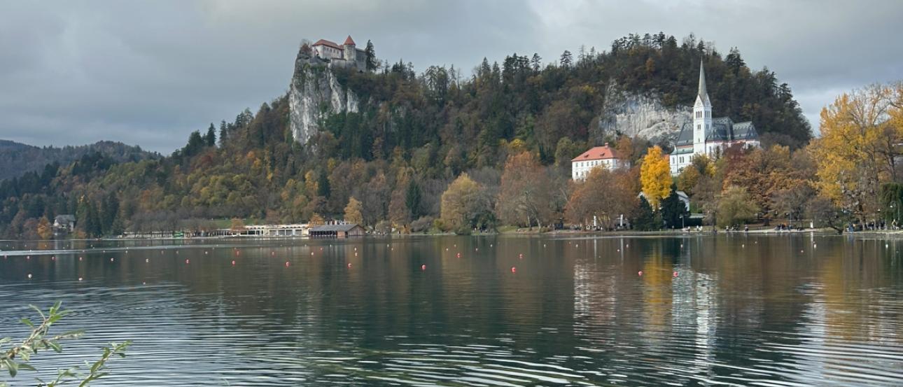 A large lake with a castle seen in the background on a large cliff with a small church at the foreground. 