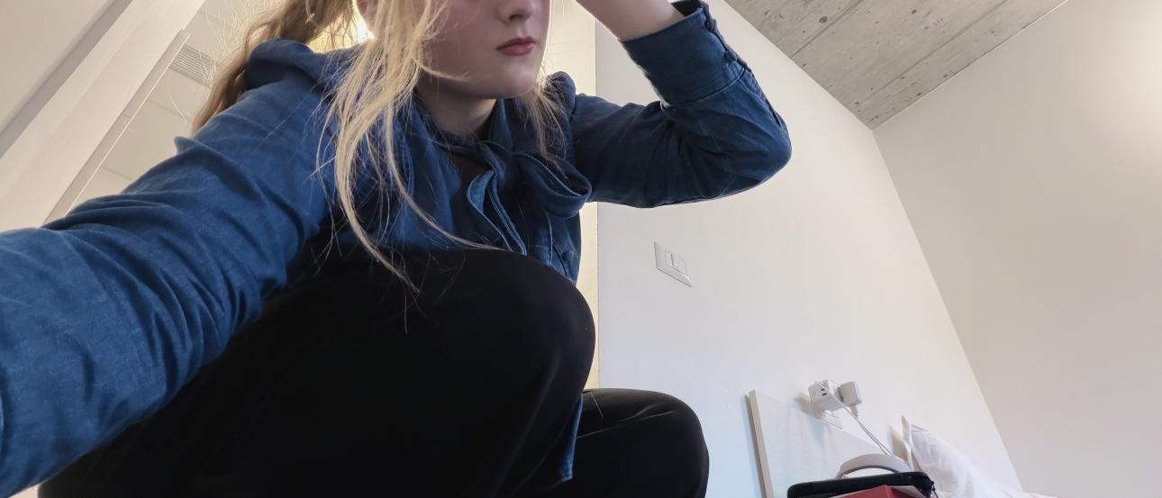 The author of the blog crouching on the floor and posing in a long sleeve denim shirt, black slacks, and white sunglasses. In the background, the Collegio dorm room is partially on display. You can see part of the grey ceiling and all white walls. 