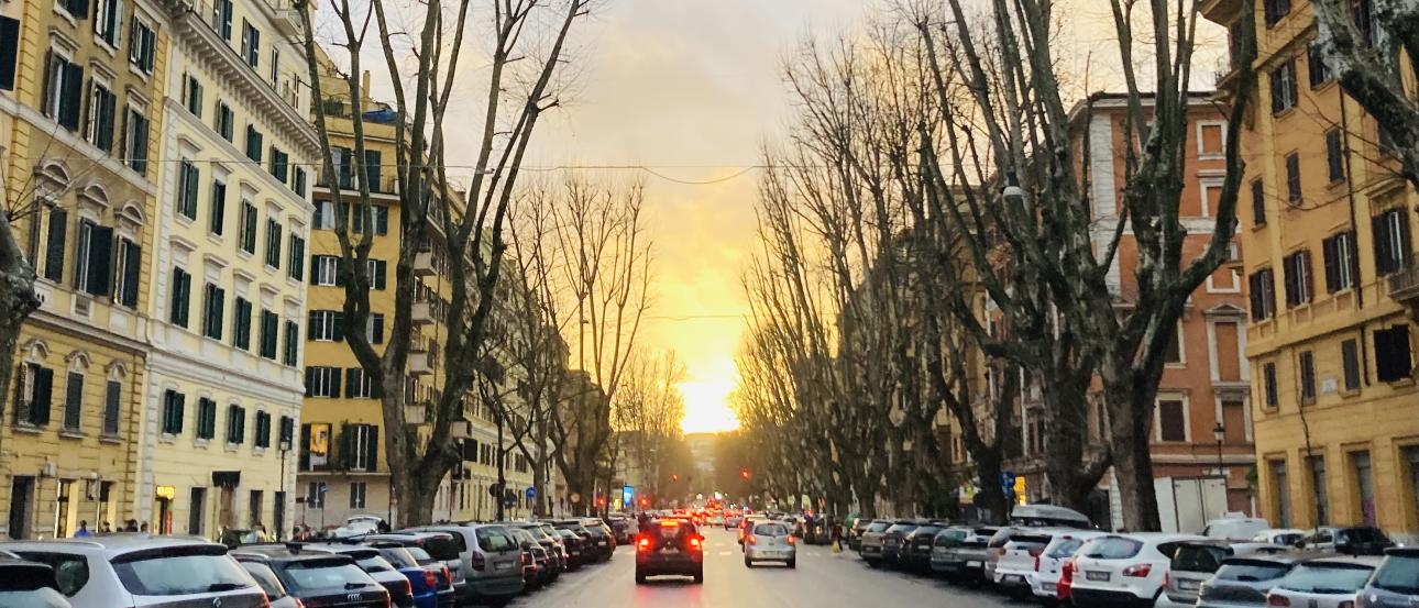 A view down a Roman street at dusk. There are cars parked on the side and the sun sets in the middle.