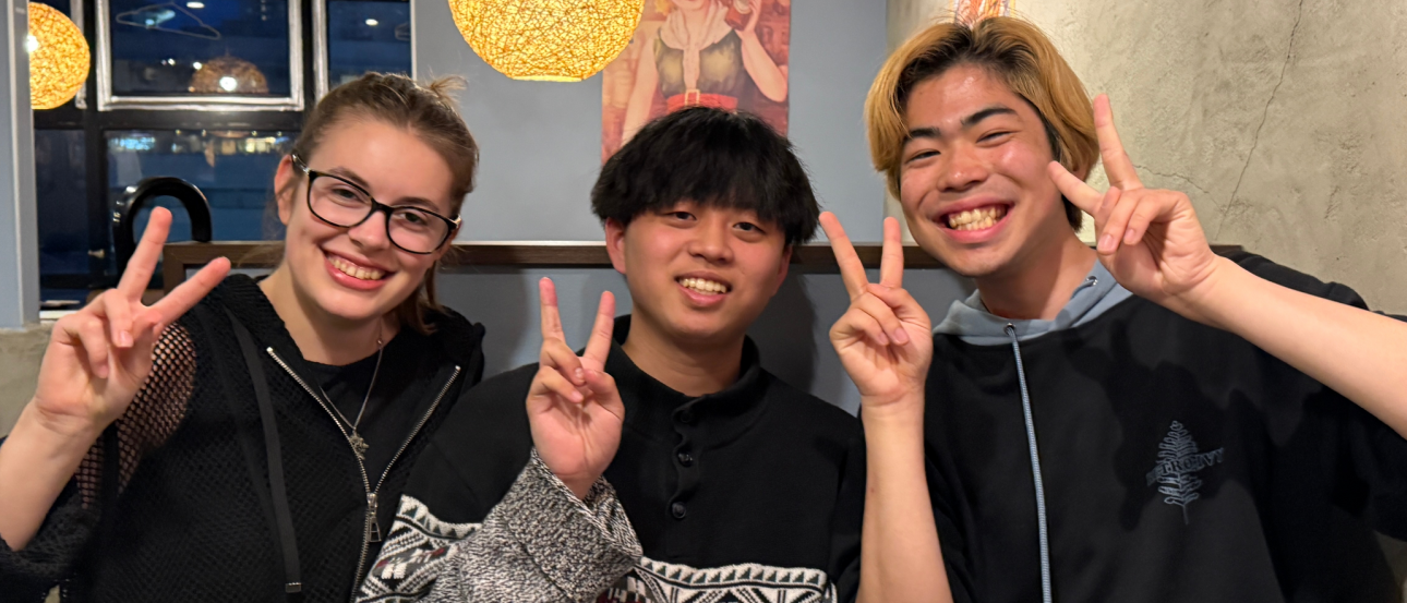 Three friends smiling and holding up peace signs while eating in a restaurant booth in Tokyo, Japan