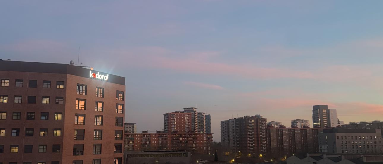 A view of the sunset through a bedroom window in Aparto Cristobal. A tall red student living building is seen along with other buildings in the area.