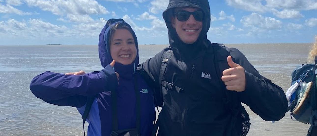 Two students smile and thumbs up for the camera, standing in the ocean