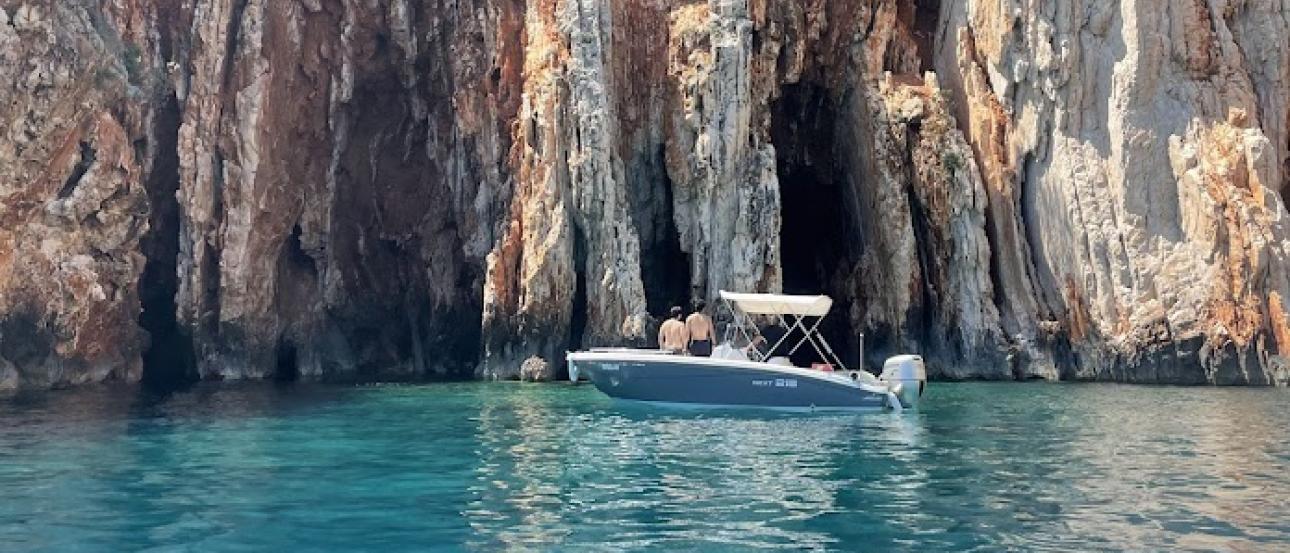 small boat floats in clear blue water and wall of reddish rocks behind it 