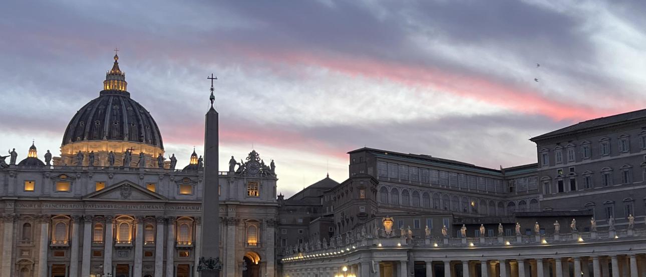 A view of St. Peter’s Basilica and St. Peter’s Square at sunset, with the basilica being illuminated by warm lights. 