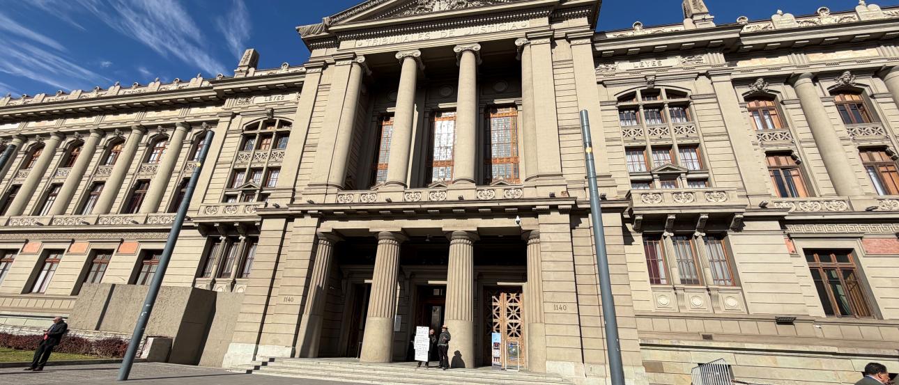 A large, tan building with pillars and detailed architecture with a Chilean flag stands against a blue sky filled with clouds. 