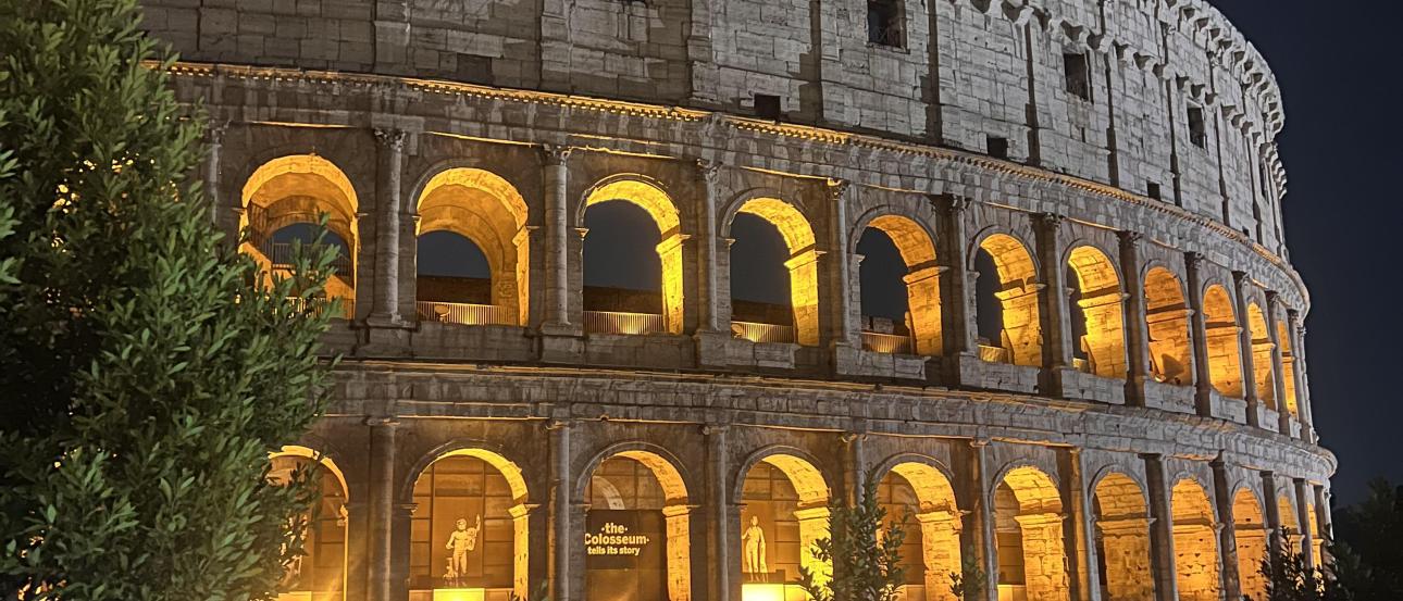  Illuminated view of the Colosseum in Rome at night, with its arches glowing yellow against the dark sky and trees in the foreground.