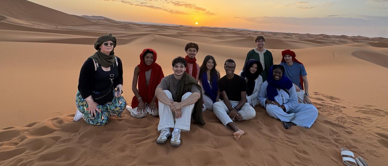 A group of students sitting on a sand dune in the Sahara Desert, Morocco. Hills of sand and a sunset is in the distance