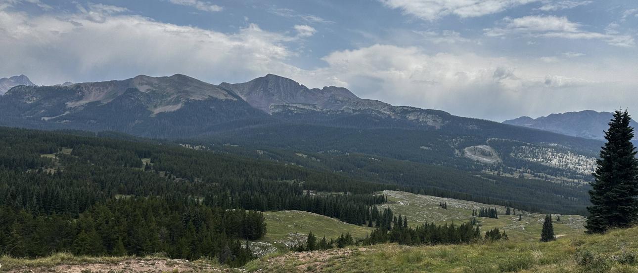 A mountain landscape, with multiple mountains and clouds in the background and greenery in the foreground.