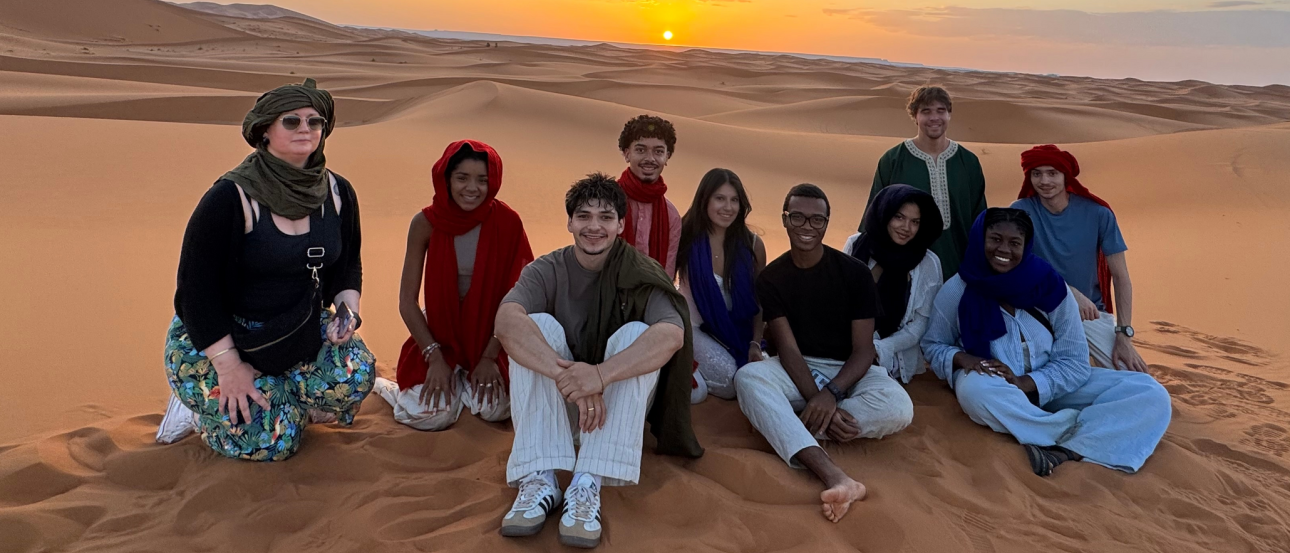 A group of students sitting on a sand dune in the Sahara Desert, Morocco. Hills of sand and a sunset is in the distance