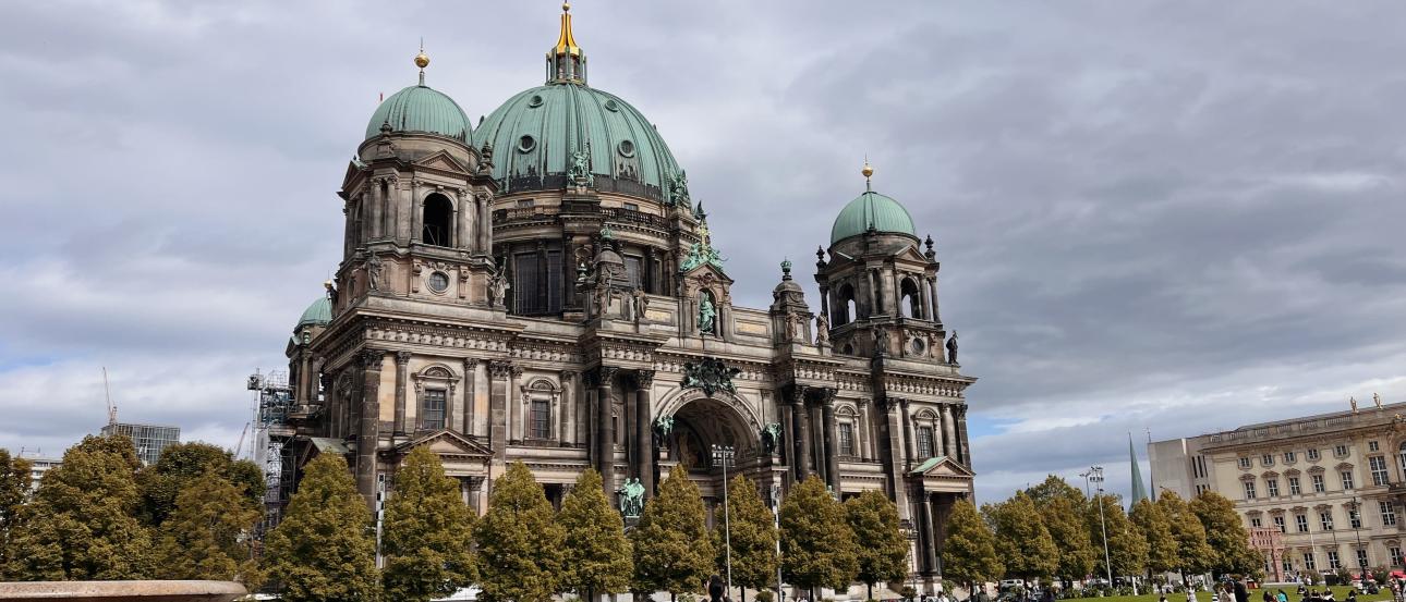 Two tourists stop to take photos in front of the Berlin Cathedral, a Baroque-style palace with a green center dome and four corner towers.