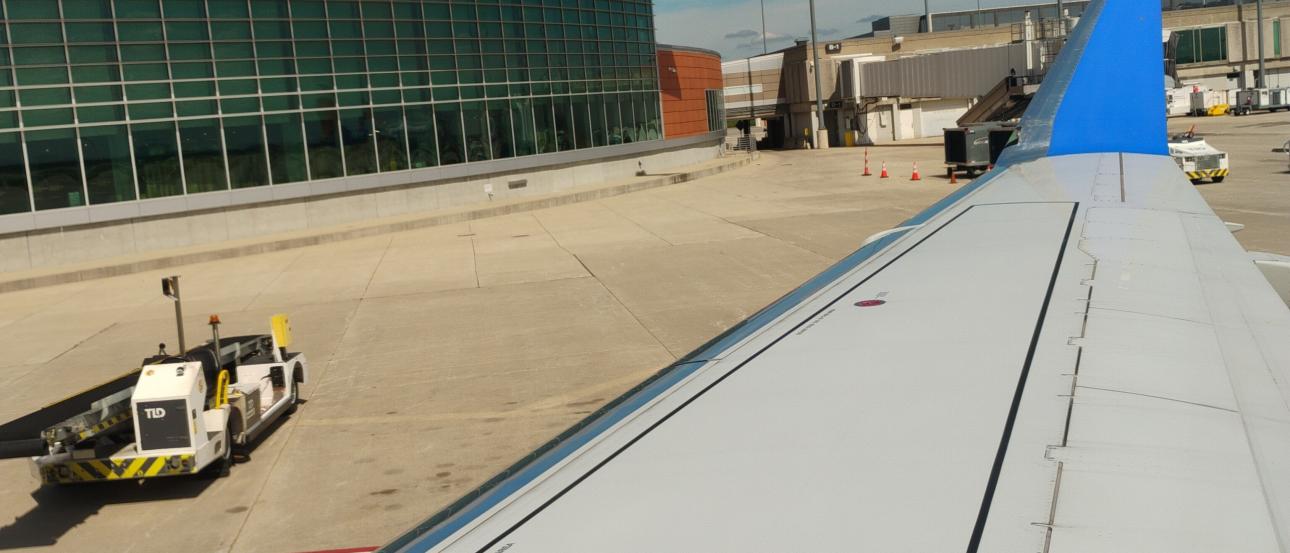 Airplane wing with a bright blue wingtip on an airport tarmac. The photo is taken from a window seat near the wing, showing the long white wing stretching toward the distance, with flap panels visible along its trailing edge. Below the wing is a concrete apron with a red guideline and some darker stained areas. Near the center-bottom of the image there is a small white and yellow ground vehicle positioned close to the wing, with a tow bar extended toward the aircraft. To the left side of the scene is an air