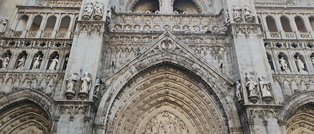 Gothic cathedral façade with a large central entrance and two side entrances, all covered in ornate stone carvings. The structure is made of pale stone and rises vertically with strong, tall pillars on the left and right sides of the main entrance. The central portal is framed by multiple pointed stone arches that recede inward, each arch ring densely filled with small sculpted figures and decorative patterns. Above the main doorway, in the tympanum area, a detailed relief scene shows many human figures.