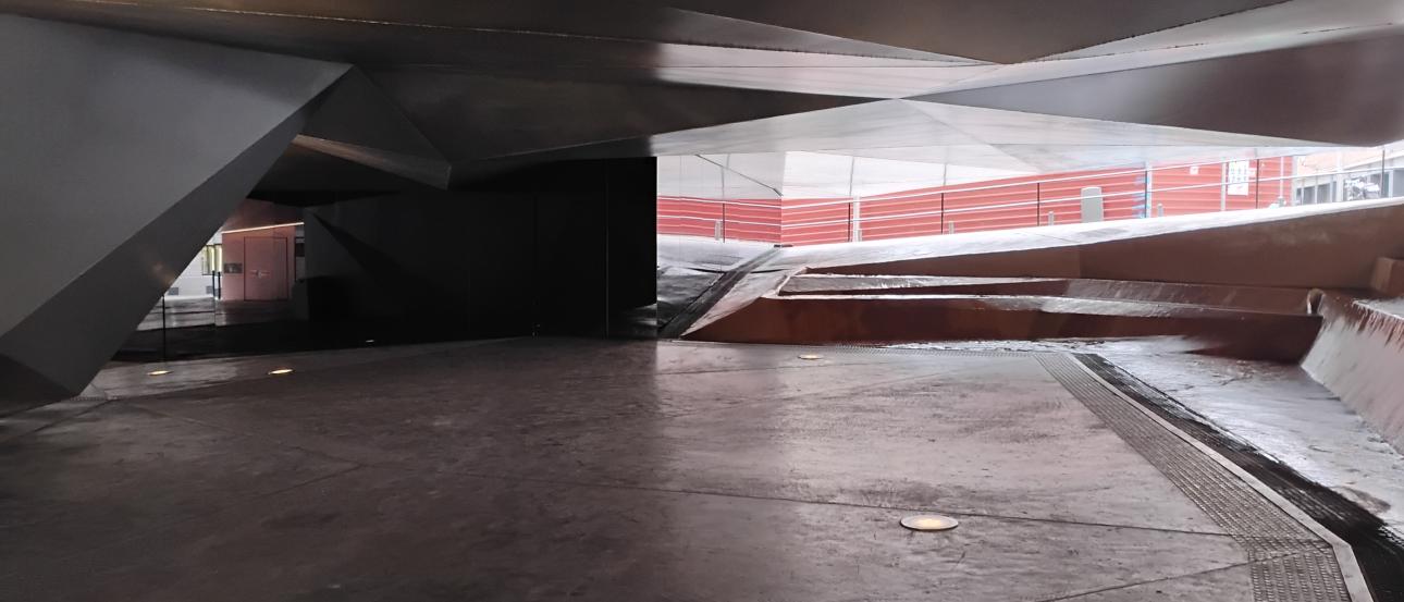 Concrete-floored underpass with angular metal ceiling panels and ramps. The image shows an interior or covered space with a smooth, slightly reflective concrete floor and several round floor lights embedded in it. The ceiling and side structures are made of large, sharp-edged metal panels forming triangular and slanted shapes, giving the space a very geometric, modern appearance. In the center-right, the floor rises in a broad ramp, with more angled surfaces leading upward toward an opening that leads outsi