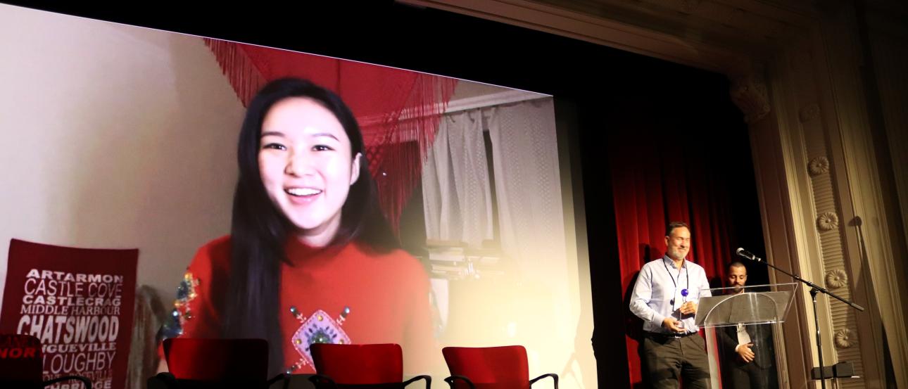 A video of Hyunseo Kong giving her acceptance speech plays on a large theater screen, while the IES Center Director holds the trophy.