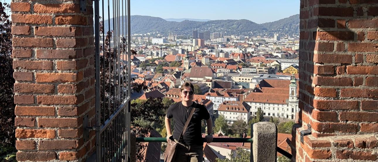 The author leans against a railing above a view of Graz, Austria, with many old buildings below. The photo is framed on both sides with brick pillars.