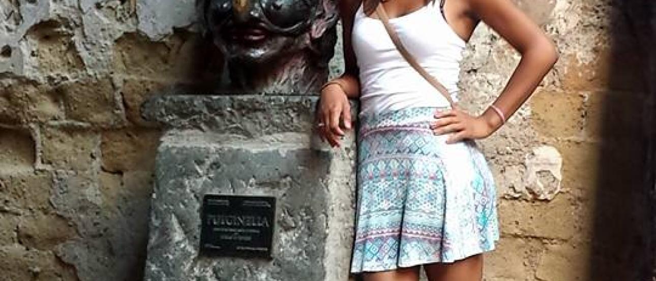 Crowd gathered on a busy street.

Standing woman in a green dress with arms outstretched in front of historic building.

Woman in a skirt next to a stone sculpture with a plaque on a narrow street. This alt text was added with Al; accuracy may vary.