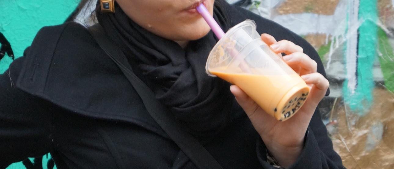 A person in a black coat drinks a beverage with graffiti in the background. This alt text was added with Al; accuracy may vary.