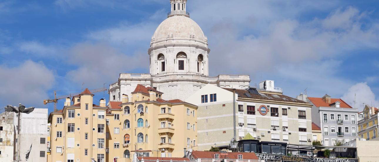 Large domed building in Lisbon with colorful houses, under a blue sky. This alt text was added with Al; accuracy may vary.