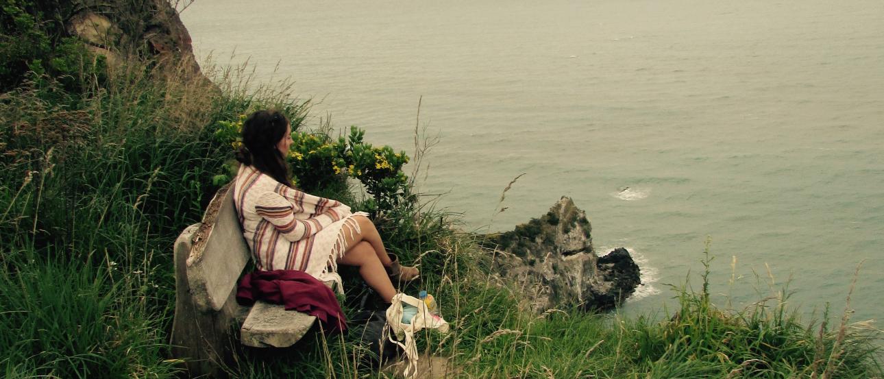 Sitting on a grassy cliff, person gazes at the ocean, overcast sky. This alt text was added with Al; accuracy may vary.