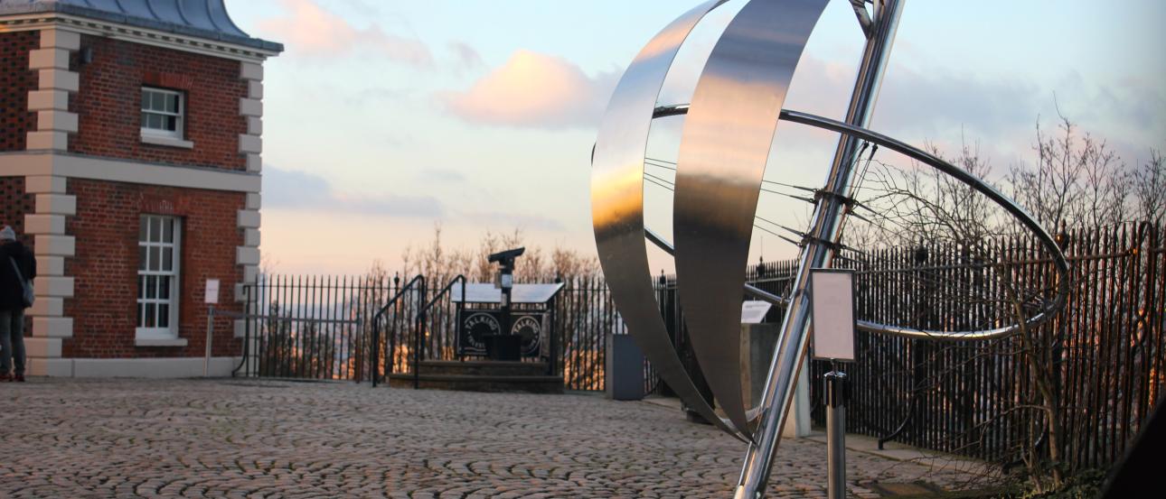 Prime Meridian sculpture at Greenwich Observatory, sunset sky in background. This alt text was added with Al; accuracy may vary.