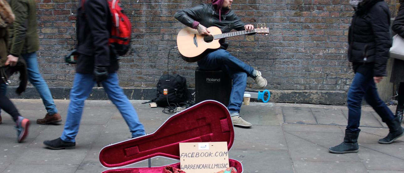 Street guitarist playing with open case on busy sidewalk. This alt text was added with Al; accuracy may vary.