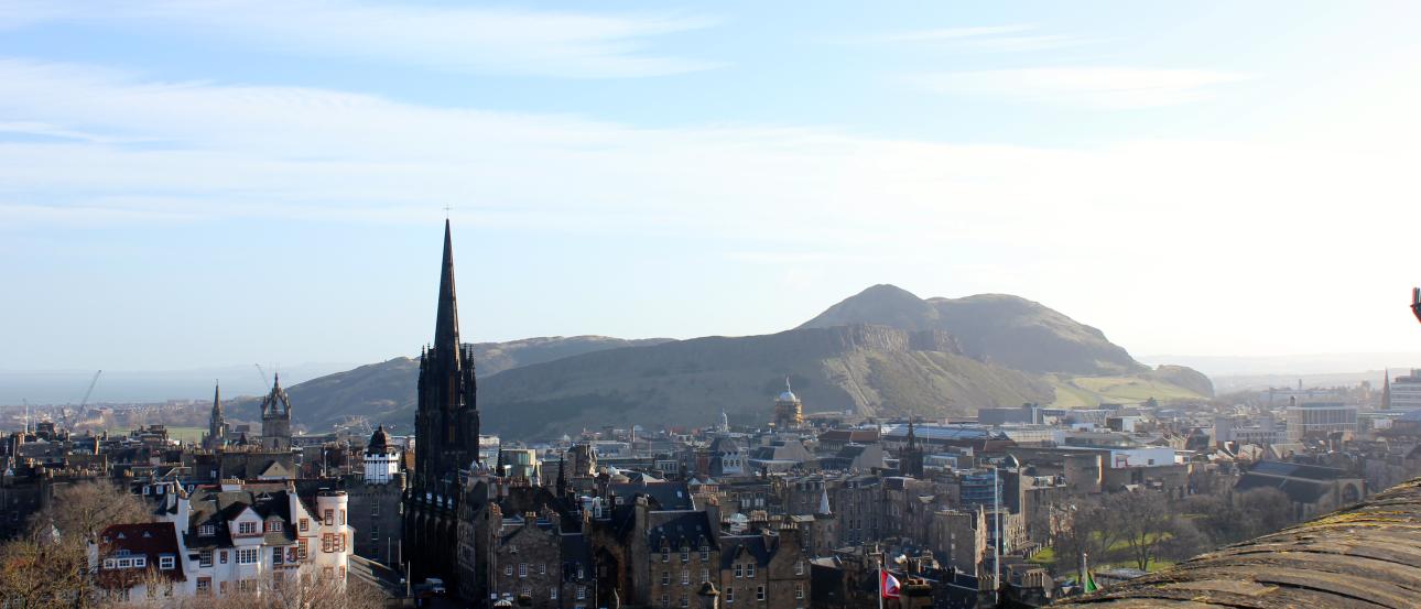 Cityscape view from a castle with a prominent spire and a hilly backdrop under a clear sky. This alt text was added with Al; accuracy may vary.