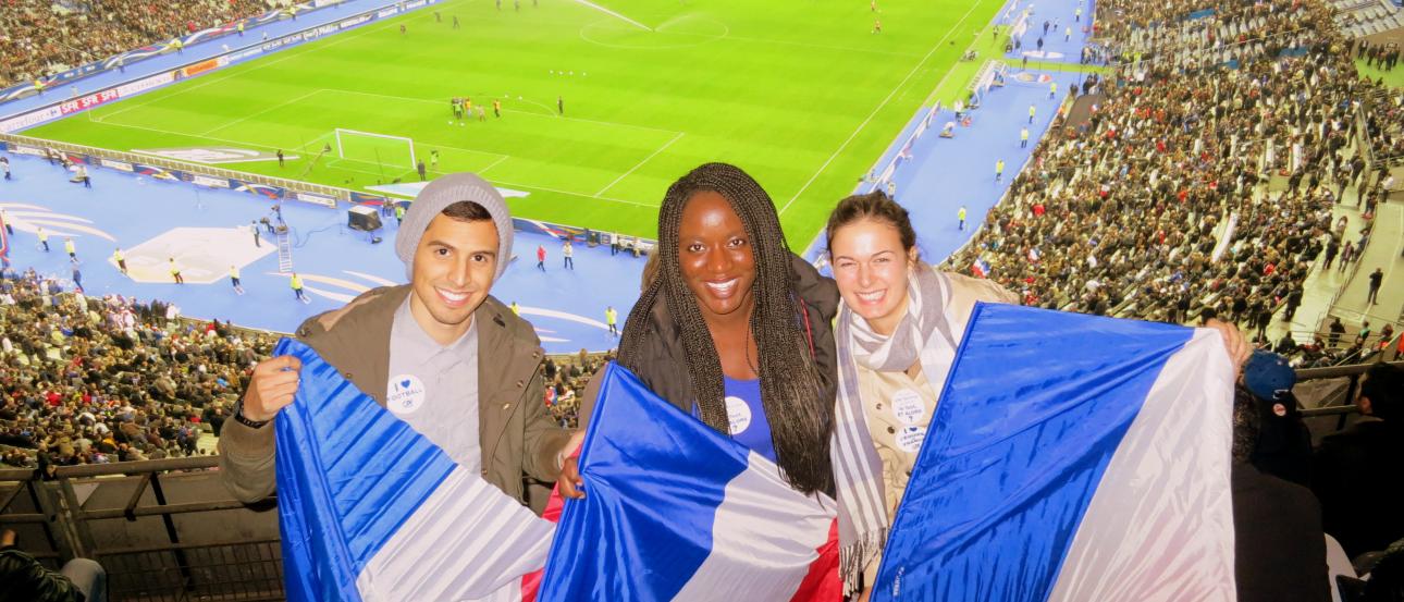 Three people holding French flags at a crowded stadium. This alt text was added with Al; accuracy may vary.