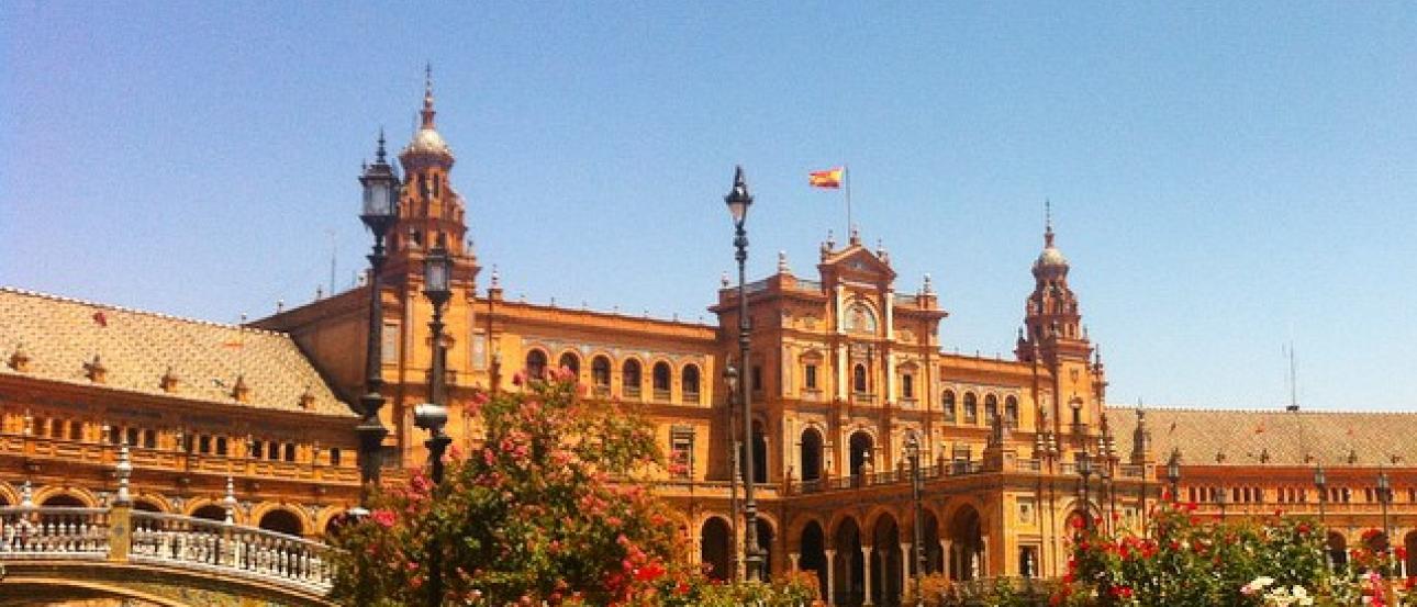 1. Tall stone church tower under cloudy sky.
2. Interior with blue stained glass and Christ figure.
3. Group of people posing before ornate archway.
4. Plaza with grand building and water canal. This alt text was added with Al; accuracy may vary.