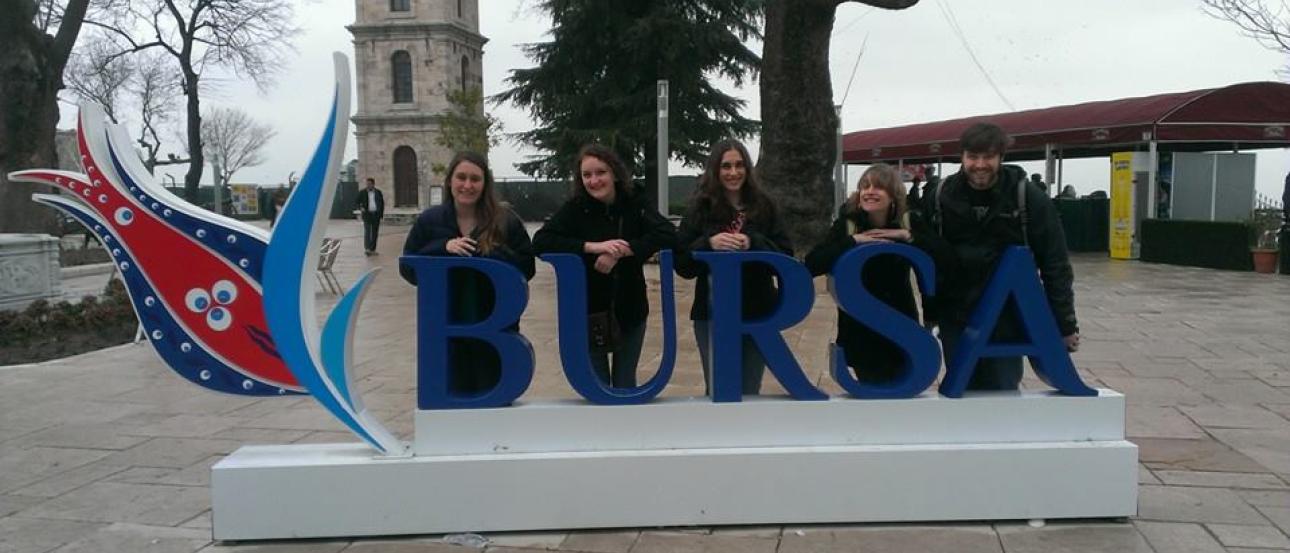 1. Cityscape with distant hills under a blue sky.
2. Colorful tulip garden near a reflective pond.
3. Messy room with cluttered items on a bed.
4. Rainbow over a lake with a castle and trees.
5. Group posing behind a "Bursa" sign in a plaza. This alt text was added with Al; accuracy may vary.