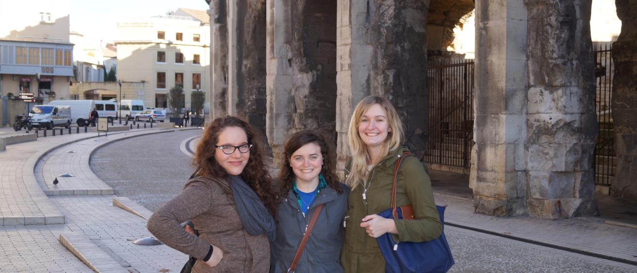 Three people smiling in front of ancient stone arches on a sunny day. This alt text was added with Al; accuracy may vary.