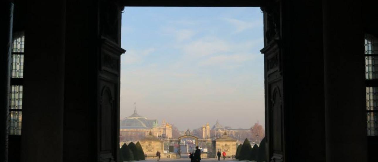 Grand archway view over hazy Paris cityscape, dominated by blue sky and distant buildings. This alt text was added with Al; accuracy may vary.