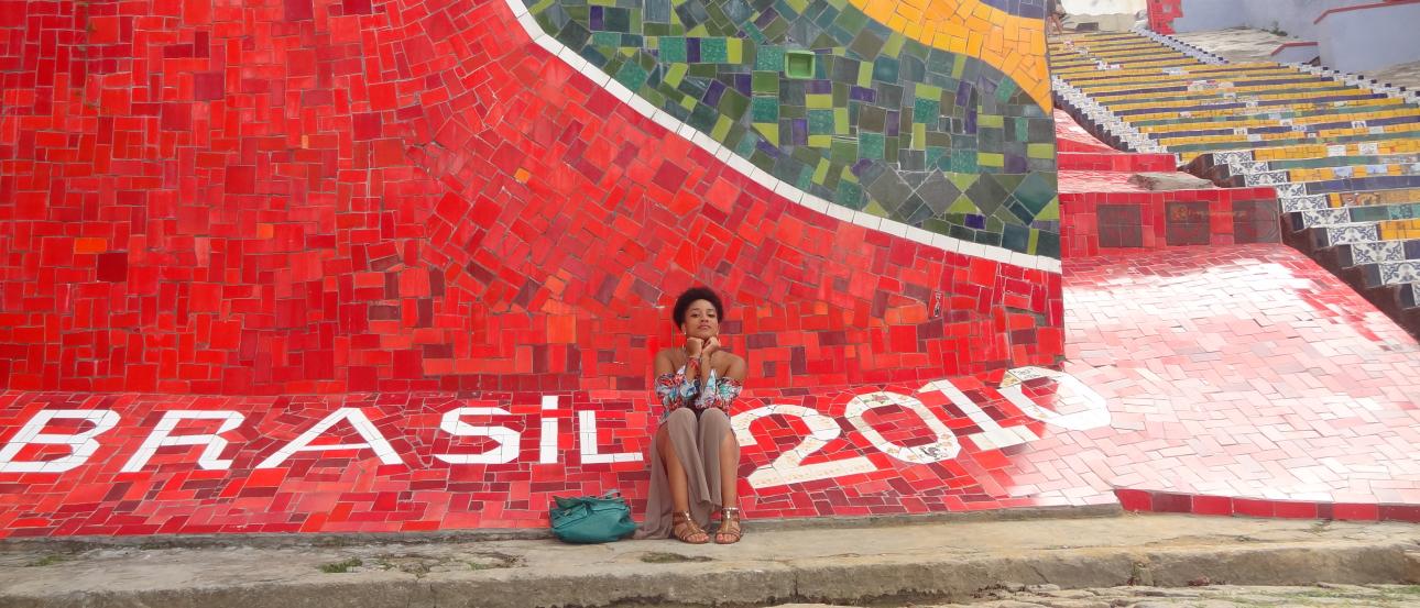 Woman sitting by colorful mosaic wall with "Brasil 2019" text. This alt text was added with Al; accuracy may vary.